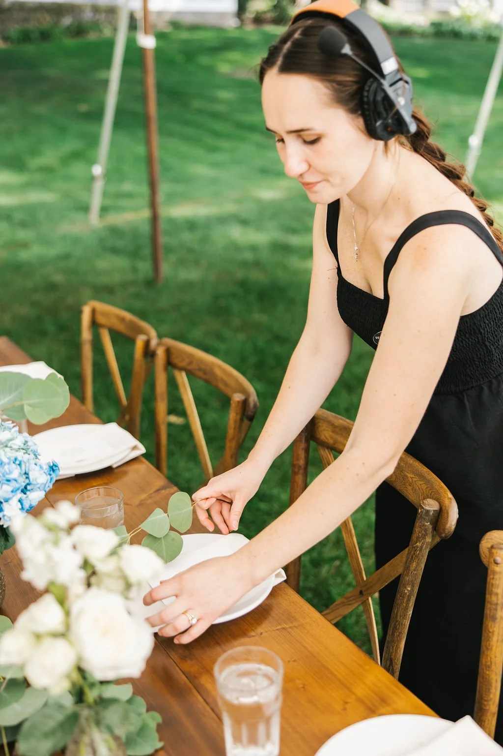 A woman wearing a black dress and a headset setting a table outdoors with white plates, a floral centerpiece, and glasses of water.