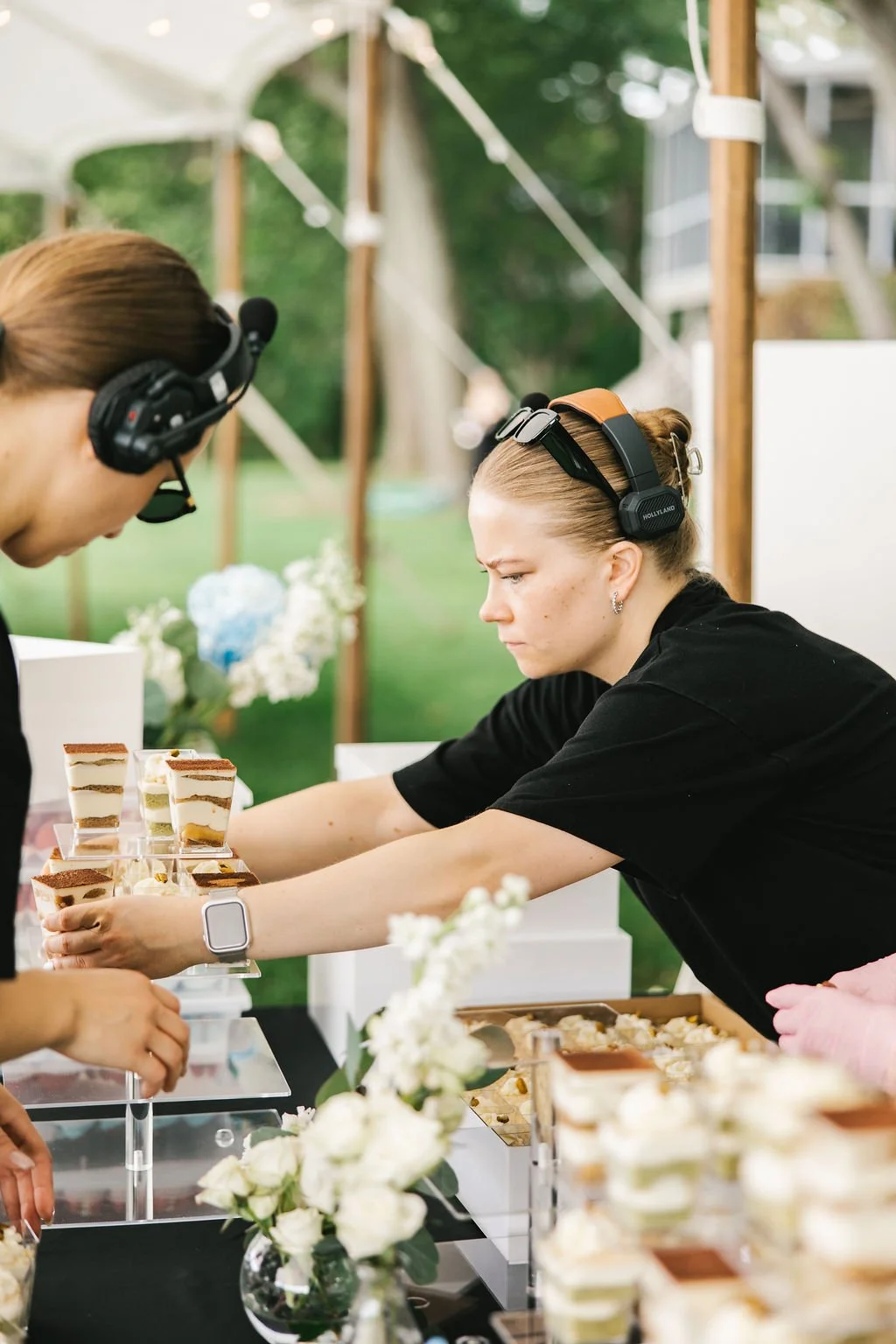 Two women wearing headsets, one with a smartwatch, at a dessert stand outdoors, arranging and selecting small layered desserts on display, with flowers and greenery in the background.