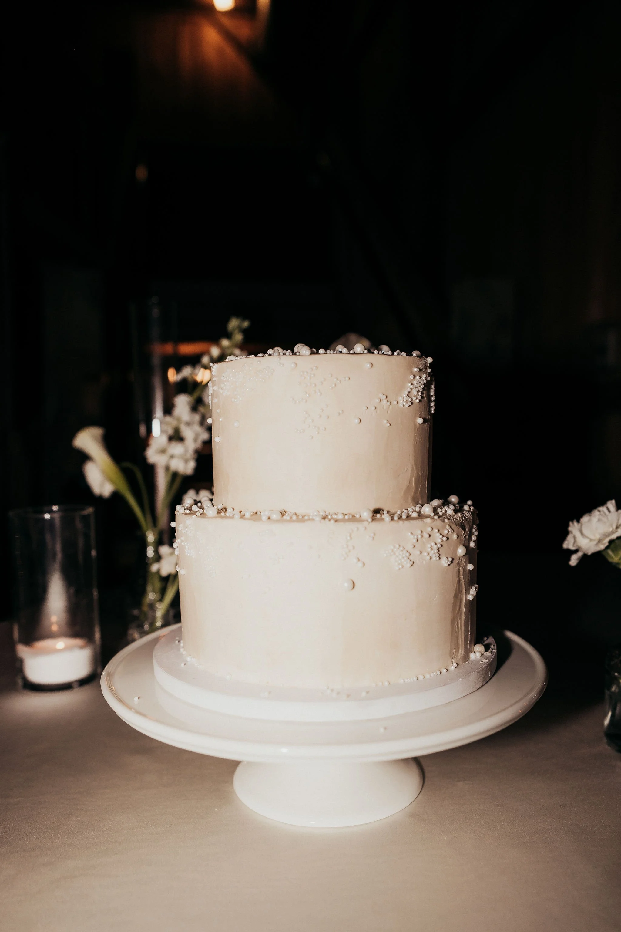 A two-tiered white wedding cake decorated with small edible pearls, placed on a white cake stand.