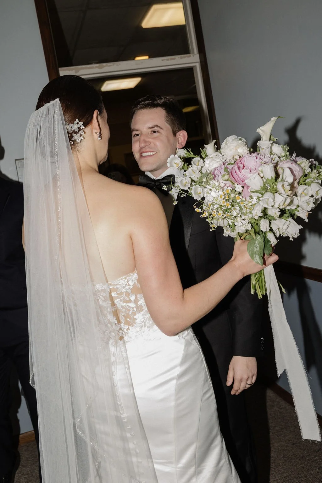 A bride and groom on their wedding day, with the bride holding a large bouquet of white and pink flowers. The bride's back is to the camera, showing her lace wedding dress and veil, while the groom is smiling at her, wearing a black tuxedo with a bow