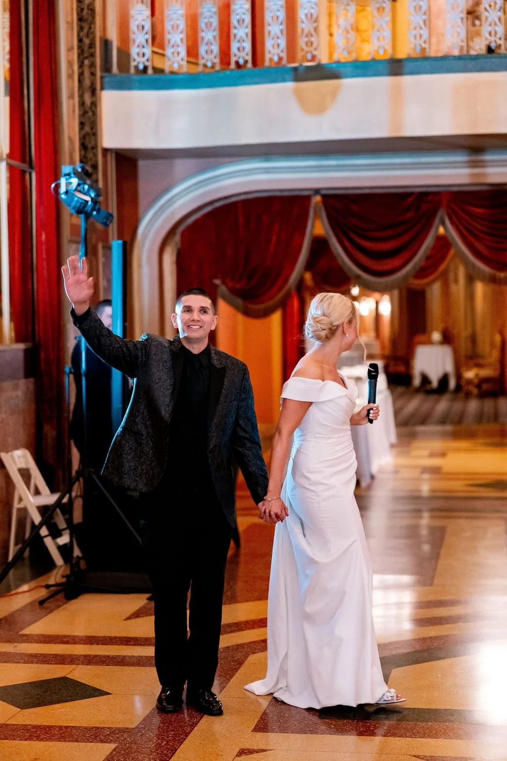 A bride and groom holding hands in a wedding reception hall. The groom is waving and smiling, wearing a black tuxedo; the bride is looking to the side, holding a microphone, wearing a white off-the-shoulder wedding gown.