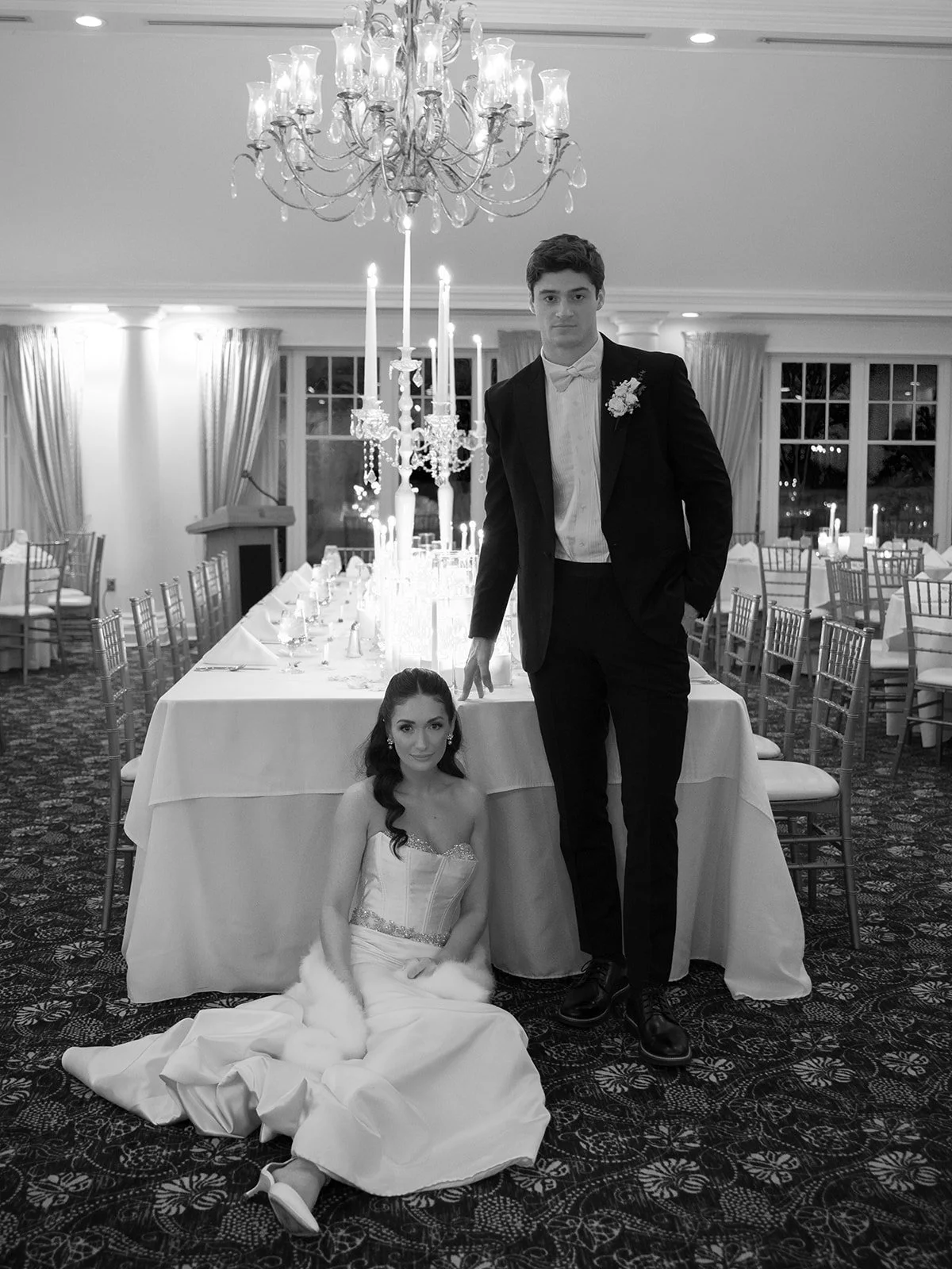 Black and white photo of a bride and groom at their wedding reception indoors. The bride is sitting on the floor in a wedding dress with a fur stole, and the groom is standing behind her with one foot on a table, wearing a tuxedo with a flower bouton