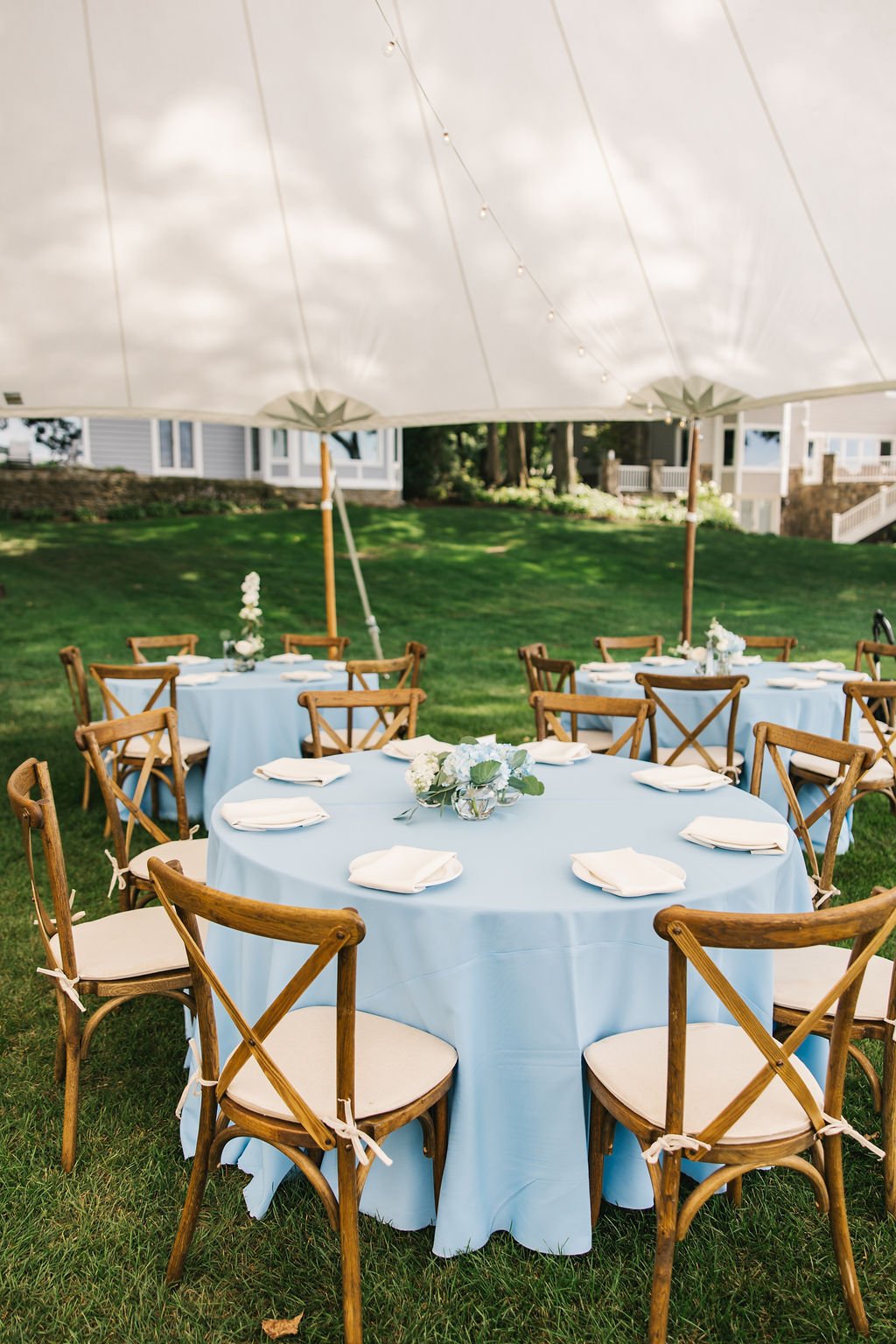 Outdoor celebration setup with round tables covered in light blue tablecloths, white napkins, and floral centerpieces, set under a large white canopy on a grassy area.