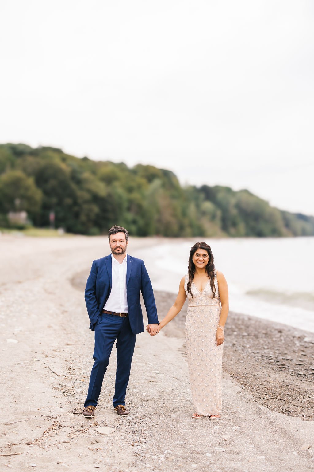 A couple holding hands on a beach, with trees and water in the background.