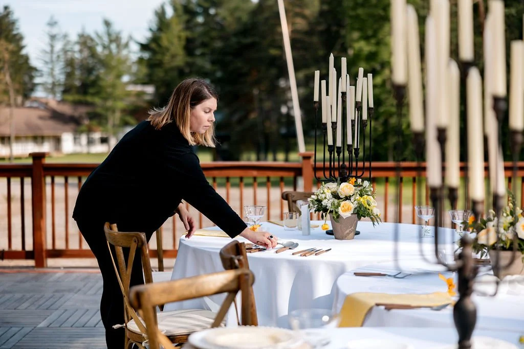 Woman setting up an outdoor dining table with floral centerpieces and candelabras on a wooden deck.