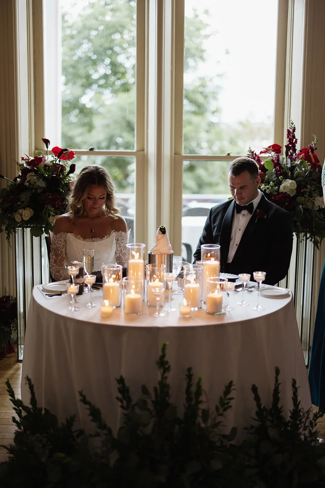 A bride and groom sitting at a round wedding reception table with lit candles, flowers, and tableware, in front of large windows with greenery outside.