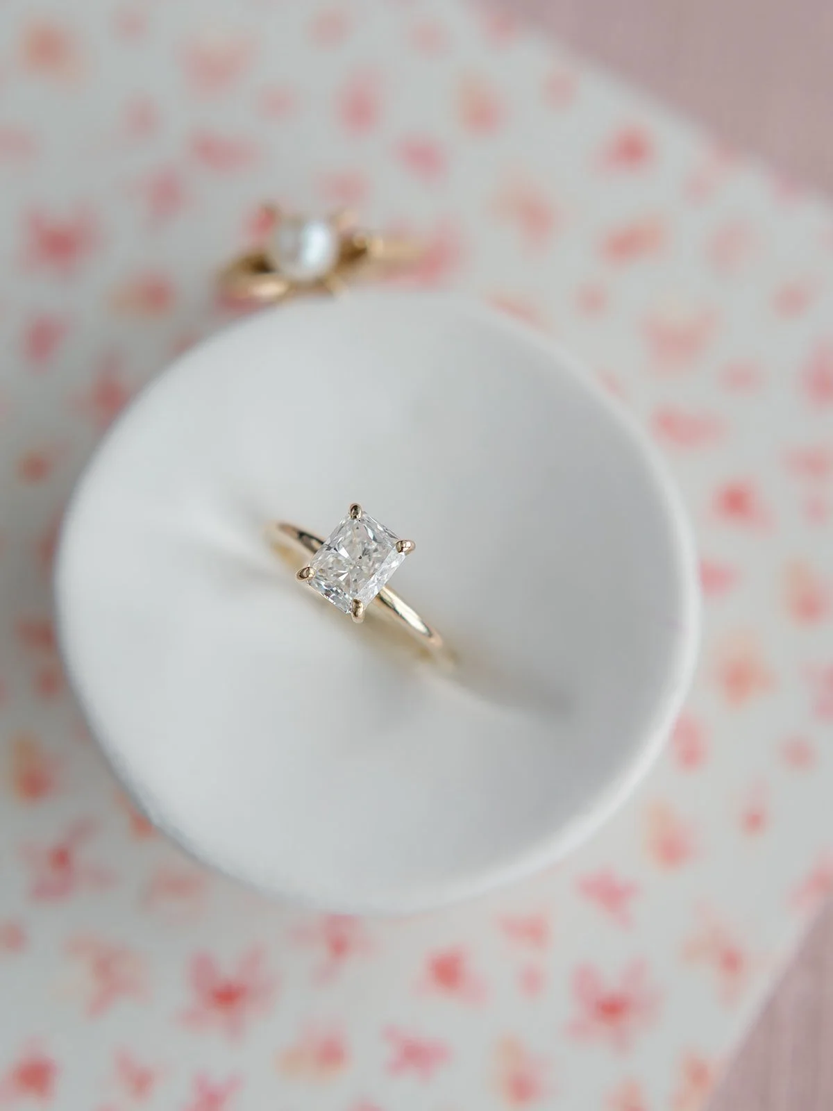 A diamond engagement ring with a square cut center stone displayed on a white ring holder, with another gold ring featuring a pearl in the background. The background has pink heart patterns.