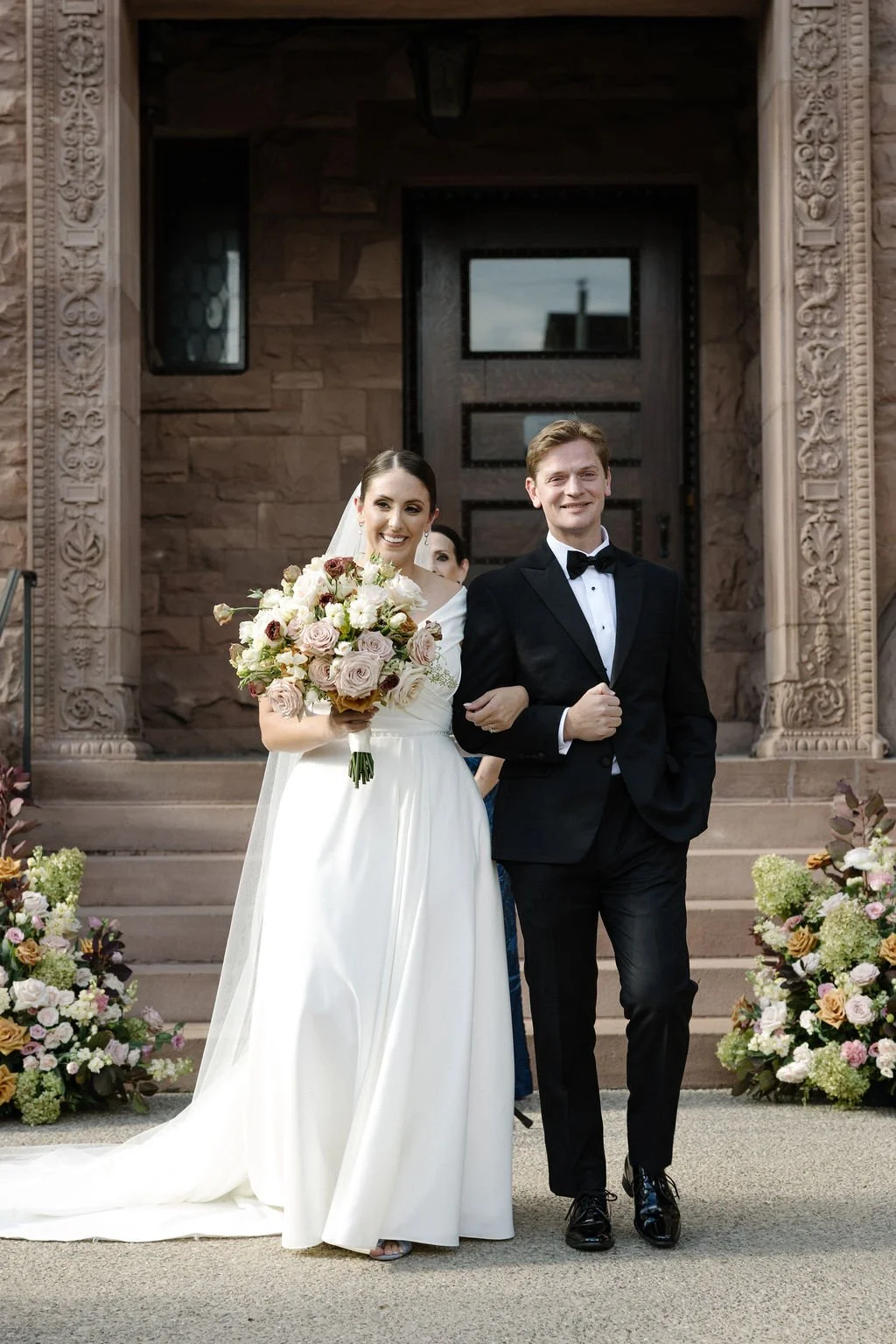Bride in a white wedding gown holding a bouquet of pink and white flowers, smiling, walking arm-in-arm with a groom in a black tuxedo and bow tie, outside a stone building decorated with floral arrangements.