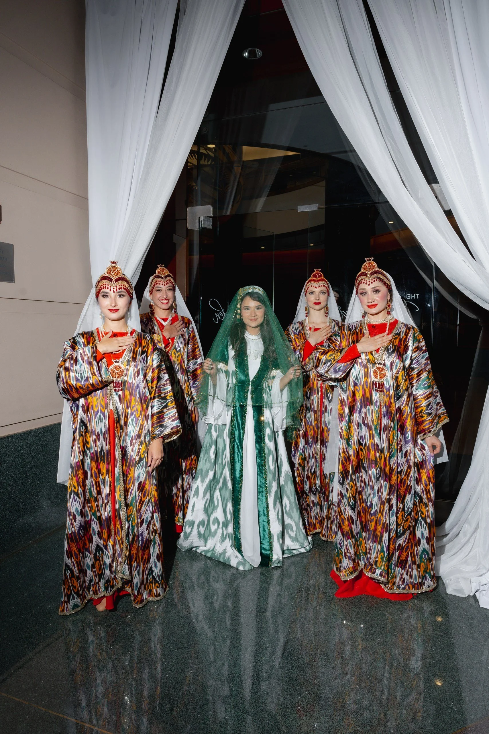 Four women dressed in traditional costumes standing in front of a glass door with white curtains. The women are wearing matching patterned dresses with red and gold accessories, and the woman in the center is wearing a white dress with green veil.
