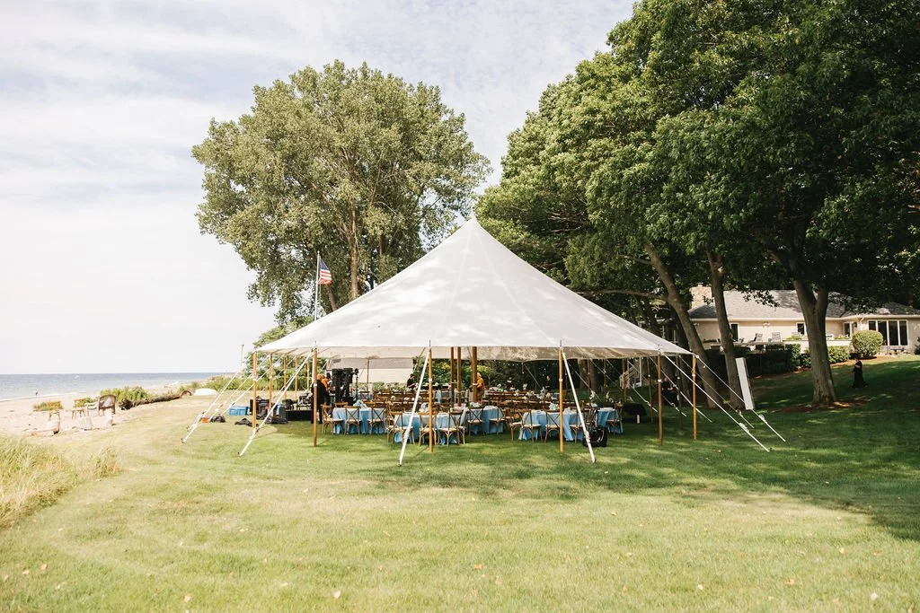 Outdoor event setup with a large white tent, decorated tables with blue tablecloths, chairs, and a view of the beach and ocean in the background, surrounded by green trees.