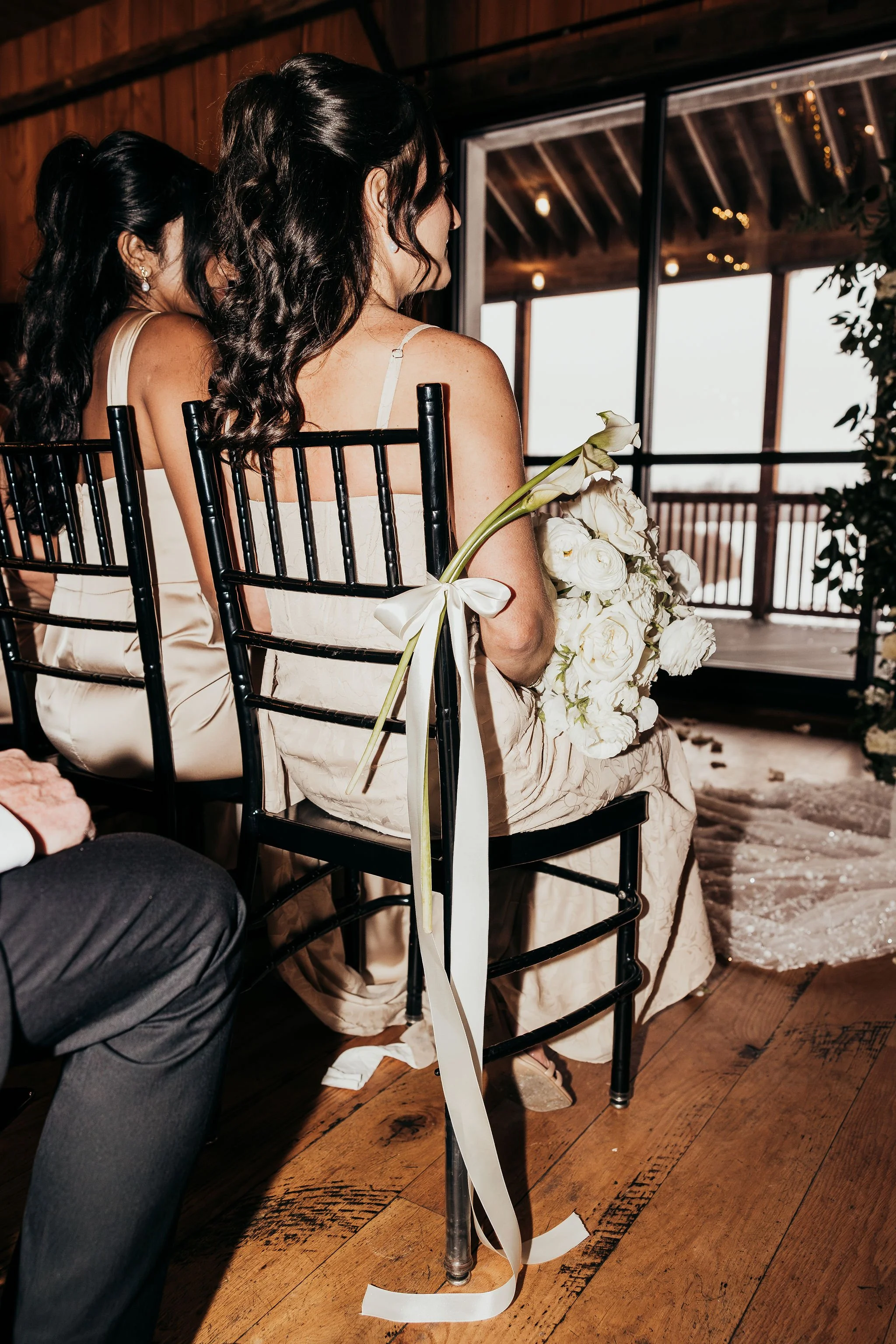 A woman with long dark wavy hair in a white dress sitting on a black chair holding a large bouquet of white flowers with a ribbon. She is at a wedding or event, with another woman partially visible beside her, in a similar dress. The setting has wood