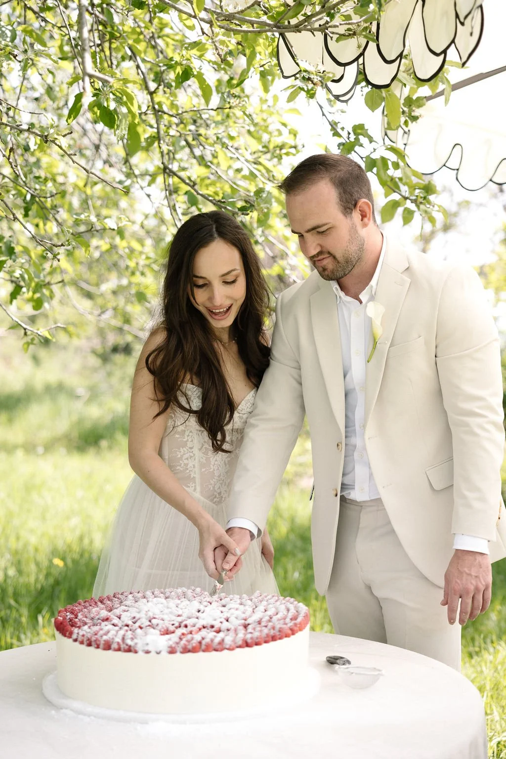 A bride and groom in wedding attire cutting a large wedding cake outdoors under a canopy of green leaves.