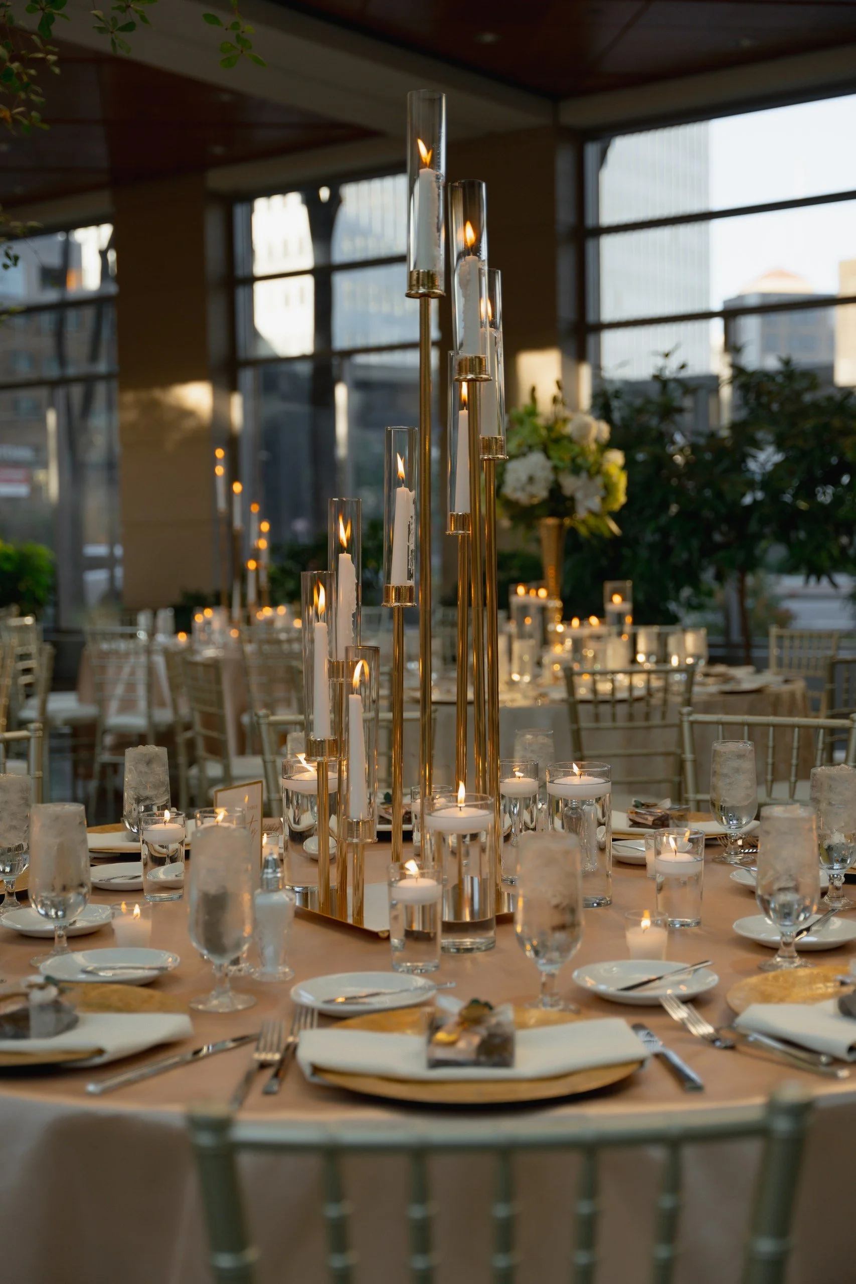 Elegant banquet table decorated with tall glass candle holders with lit candles, surrounded by water glasses, plates, silverware, and white napkins, with floral arrangements in the background, set in a venue with large windows.