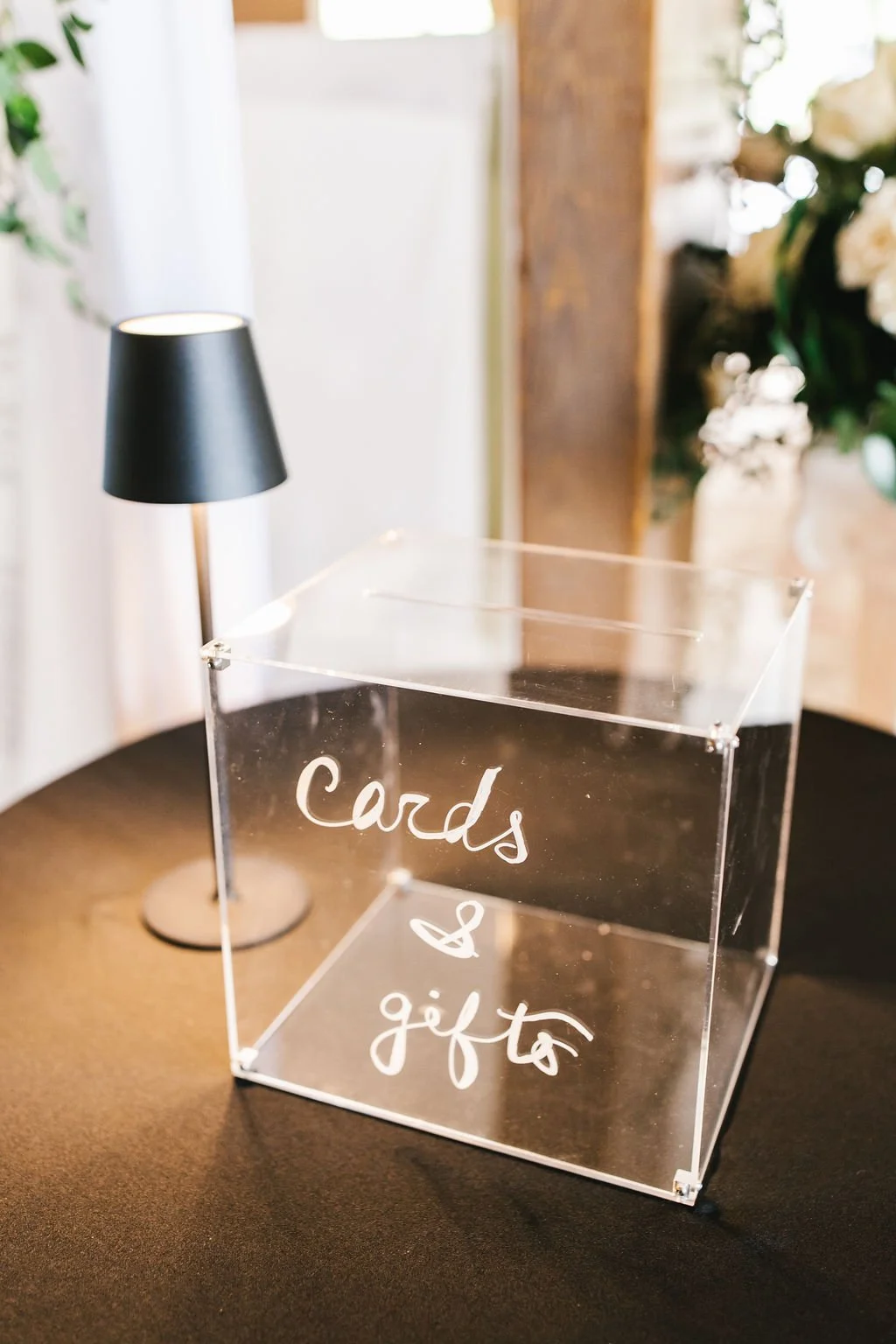 A clear acrylic box with the words 'cards & gifts' written in white on the front, sitting on a black table next to a small black table lamp with a black shade.