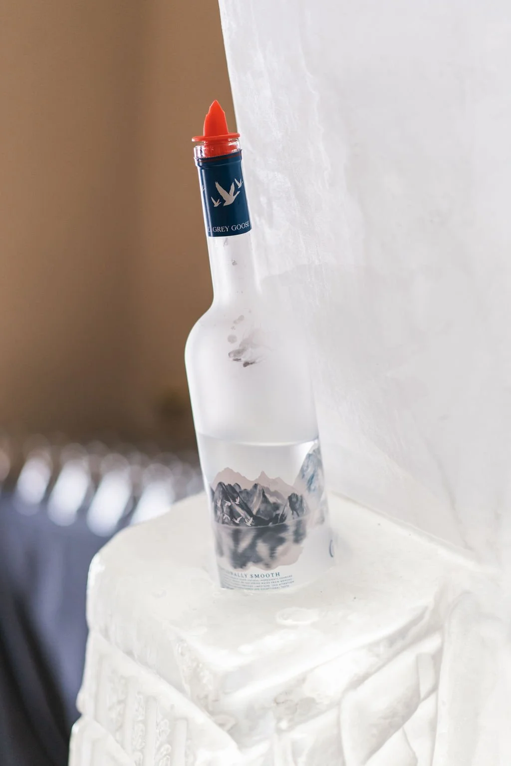 A decorative glass bottle with mountain artwork and a red lipstick-shaped cap, placed on a white surface next to a white cloth.