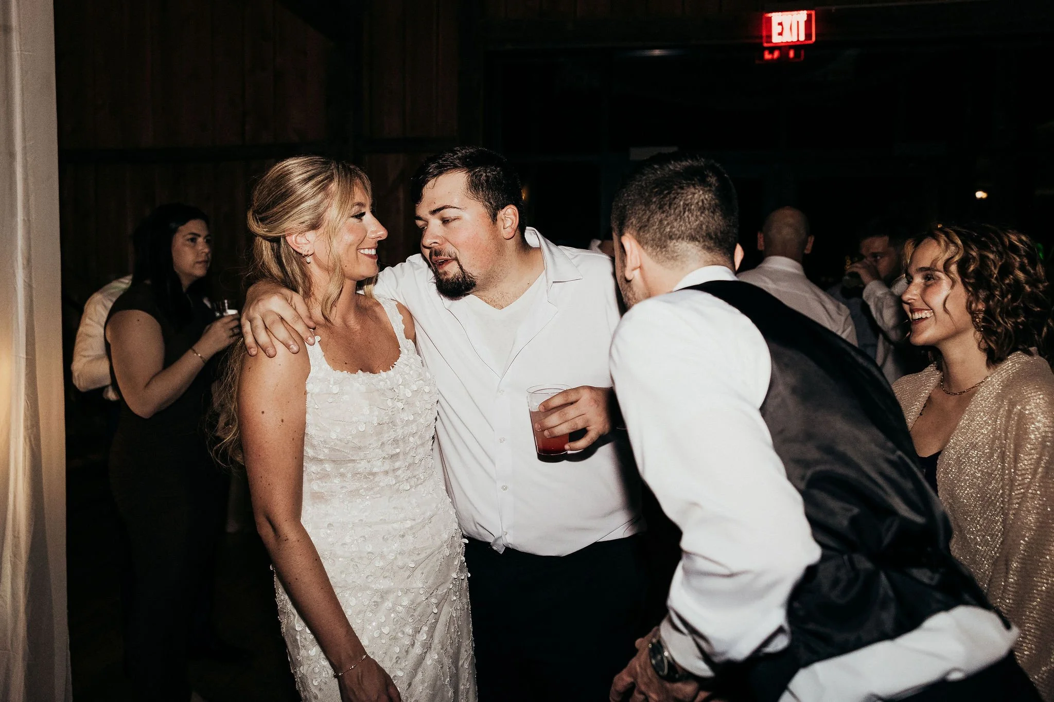 People at a wedding reception, including a bride in a white lace dress, smiling and talking with guests in a dimly lit venue.