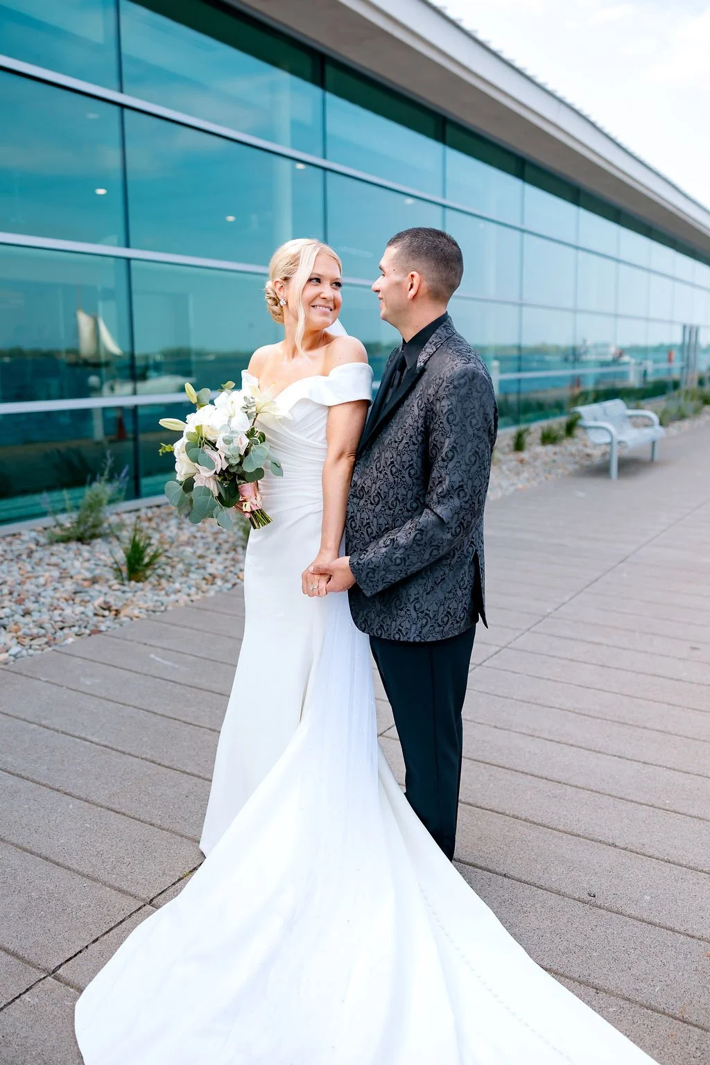 A bride and groom holding hands and smiling at each other on an outdoor walkway with a modern glass building in the background.