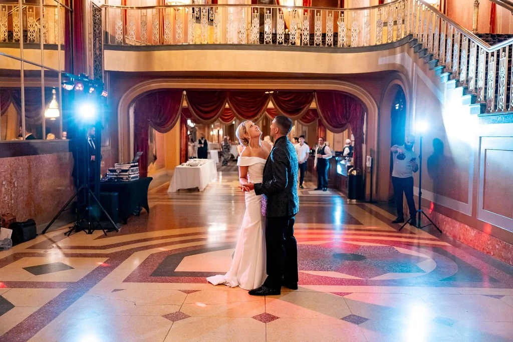 Bride and groom sharing a dance in a grand, elegant hall with wooden floors, ornate staircase, and red velvet curtains.