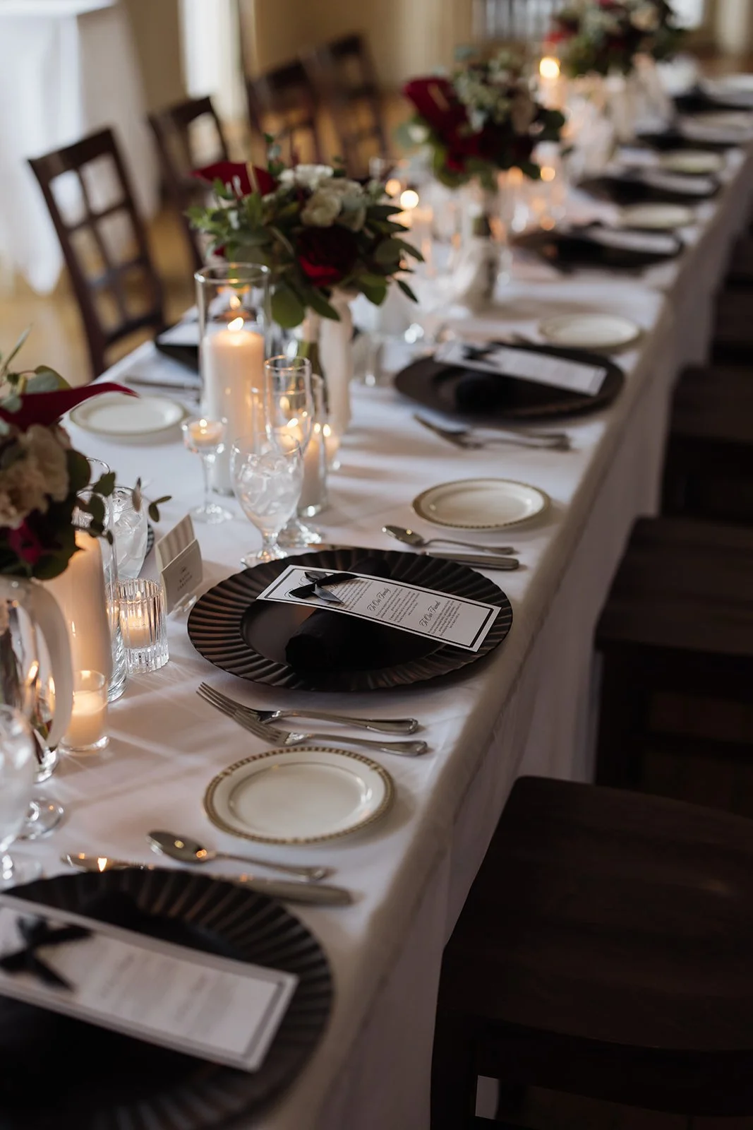 A long dining table set for a formal event with floral centerpieces, candles, black and gold plates, and silverware.