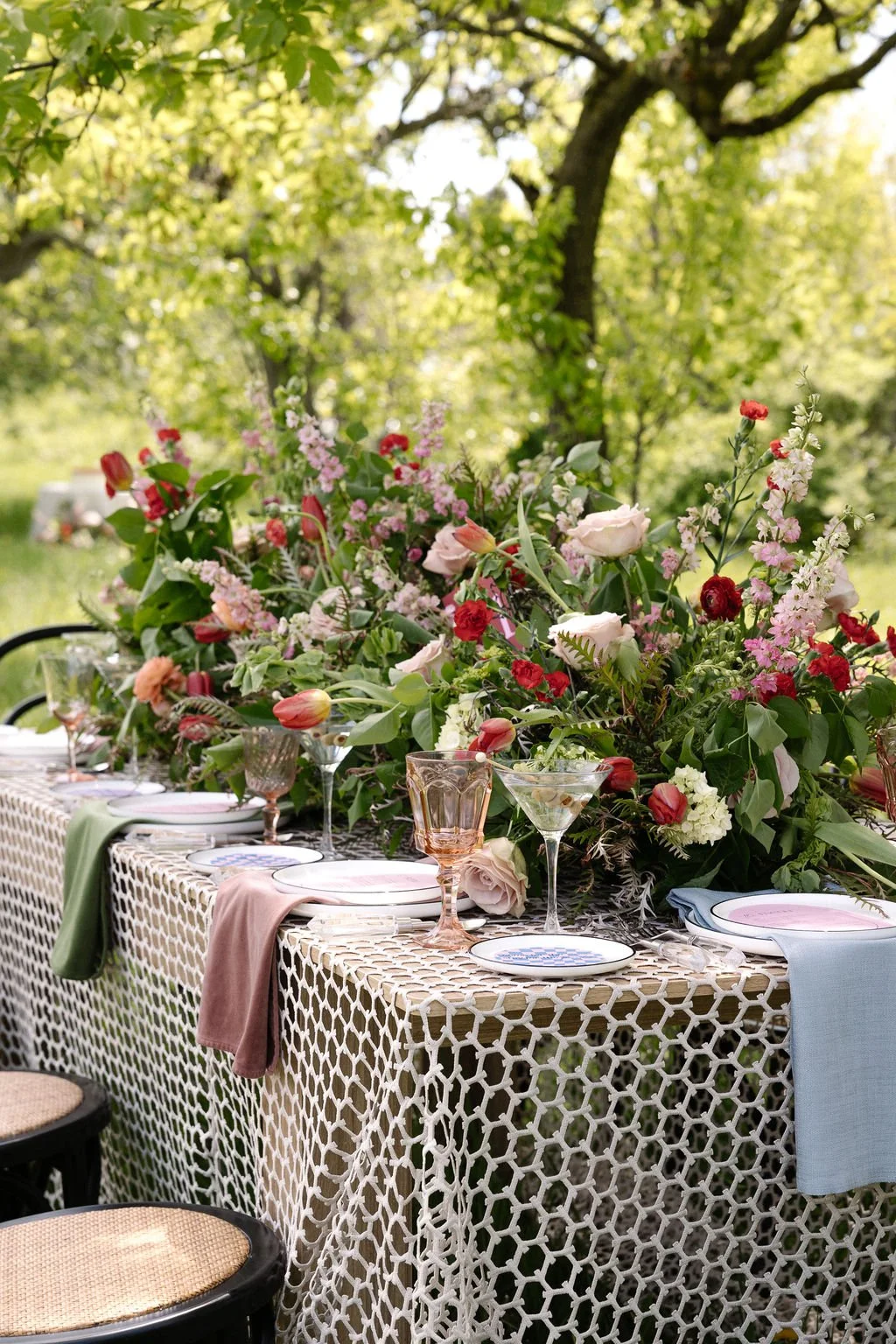 Outdoor arranged dining table with a large floral centerpiece, pastel-colored napkins, and glassware, set in a lush green garden with trees.