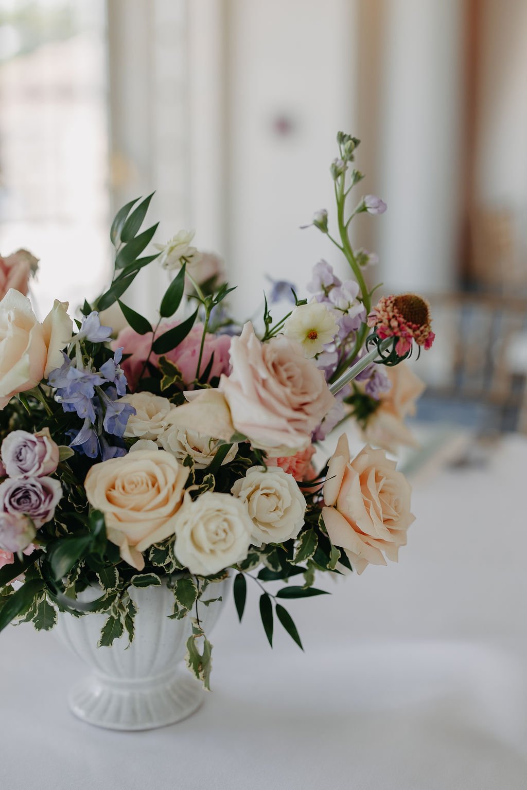 A floral arrangement with light pink, cream, and purple roses, along with green and purple filler flowers, in a white vase on a white surface.