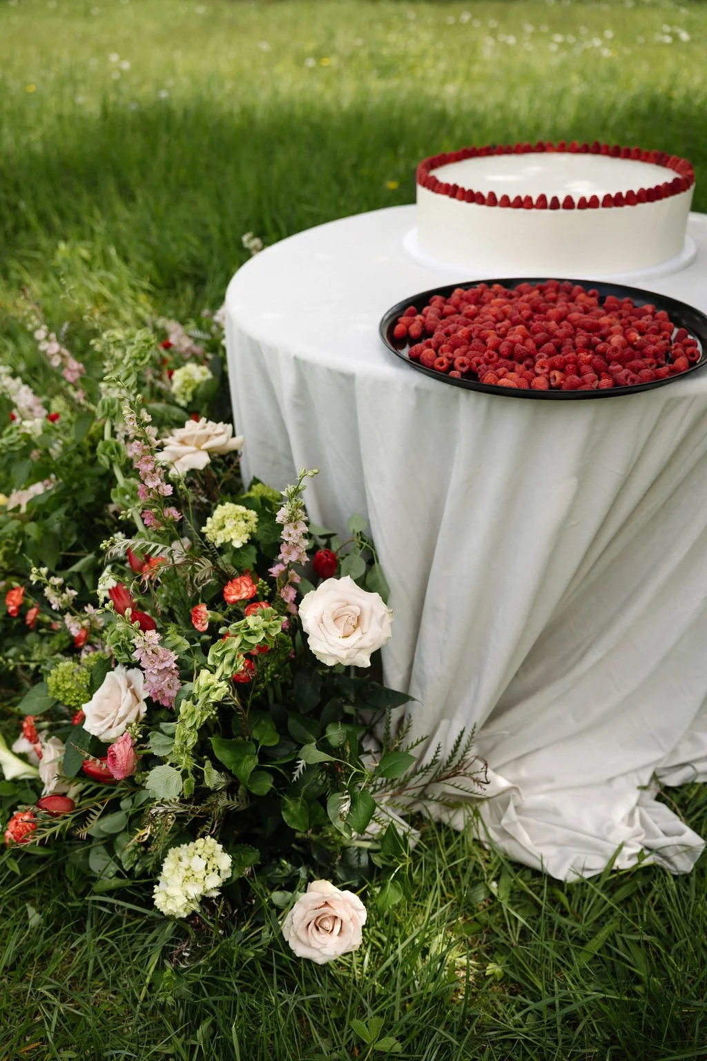 A white cake with red berries decoration on top, placed on a round table with a white cloth, outdoors on green grass garden, next to a floral arrangement with roses, small blooms, and green leaves.