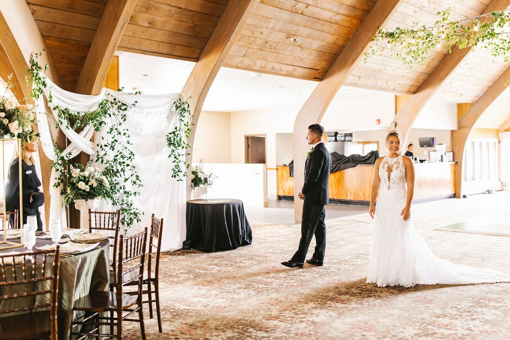 Bride in a white lace wedding gown smiling, groom in a dark suit standing nearby inside a decorated wedding venue with wooden beams and floral arrangements.