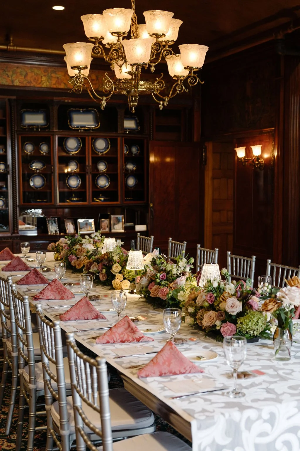 Elegant dining room with a long table decorated with floral arrangements, pink napkins, and glassware, illuminated by a chandelier and wall sconces, with a dark wooden bookcase displaying plates and framed photographs in the background.