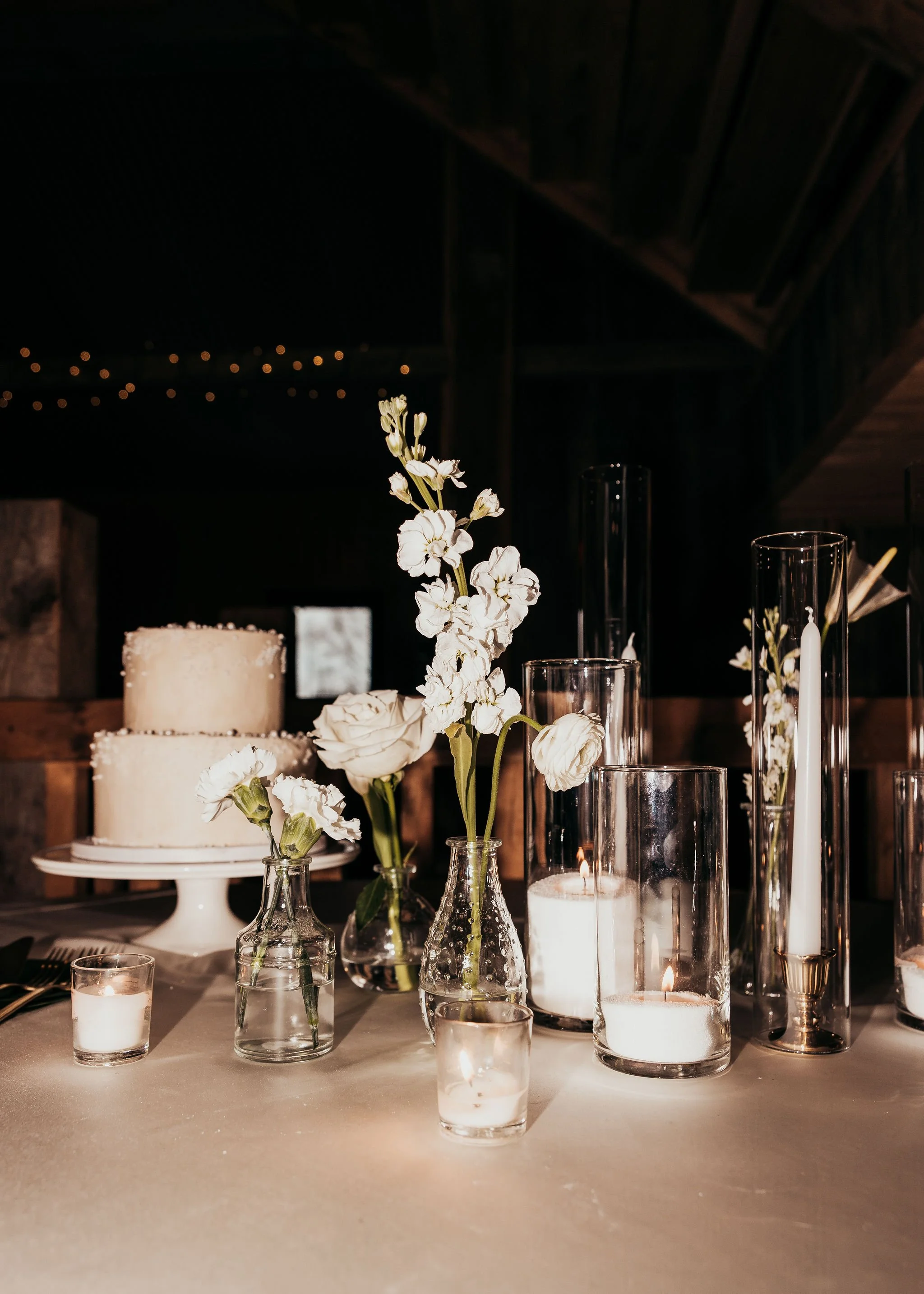 Elegant table centerpiece with white flowers in glass vases, surrounded by candles in various glass holders, with a layered white cake in the background, against a dark setting with string lights overhead.