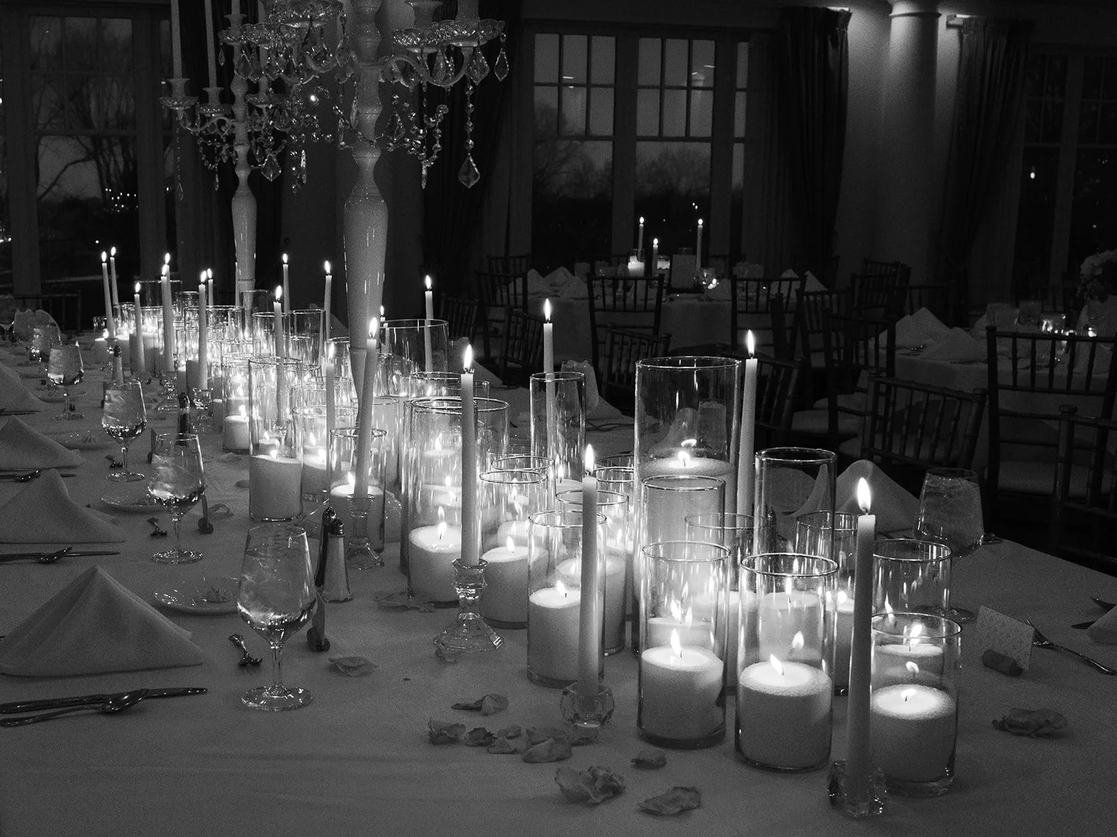 A black and white photo of a dining table decorated with numerous lit candles in glass and candle holders, with folded napkins and wine glasses set around it. A crystal chandelier hangs above, and large windows with curtains are in the background, su