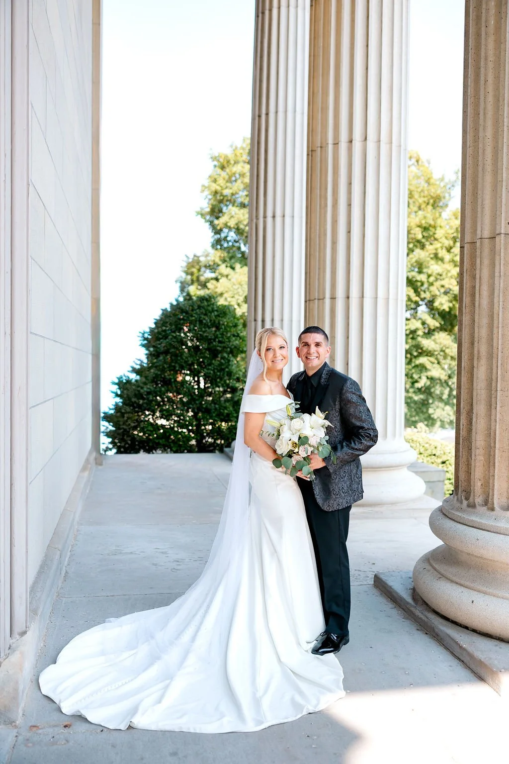 A bride and groom standing outside a classical building with tall columns, smiling and holding a bouquet of white flowers.