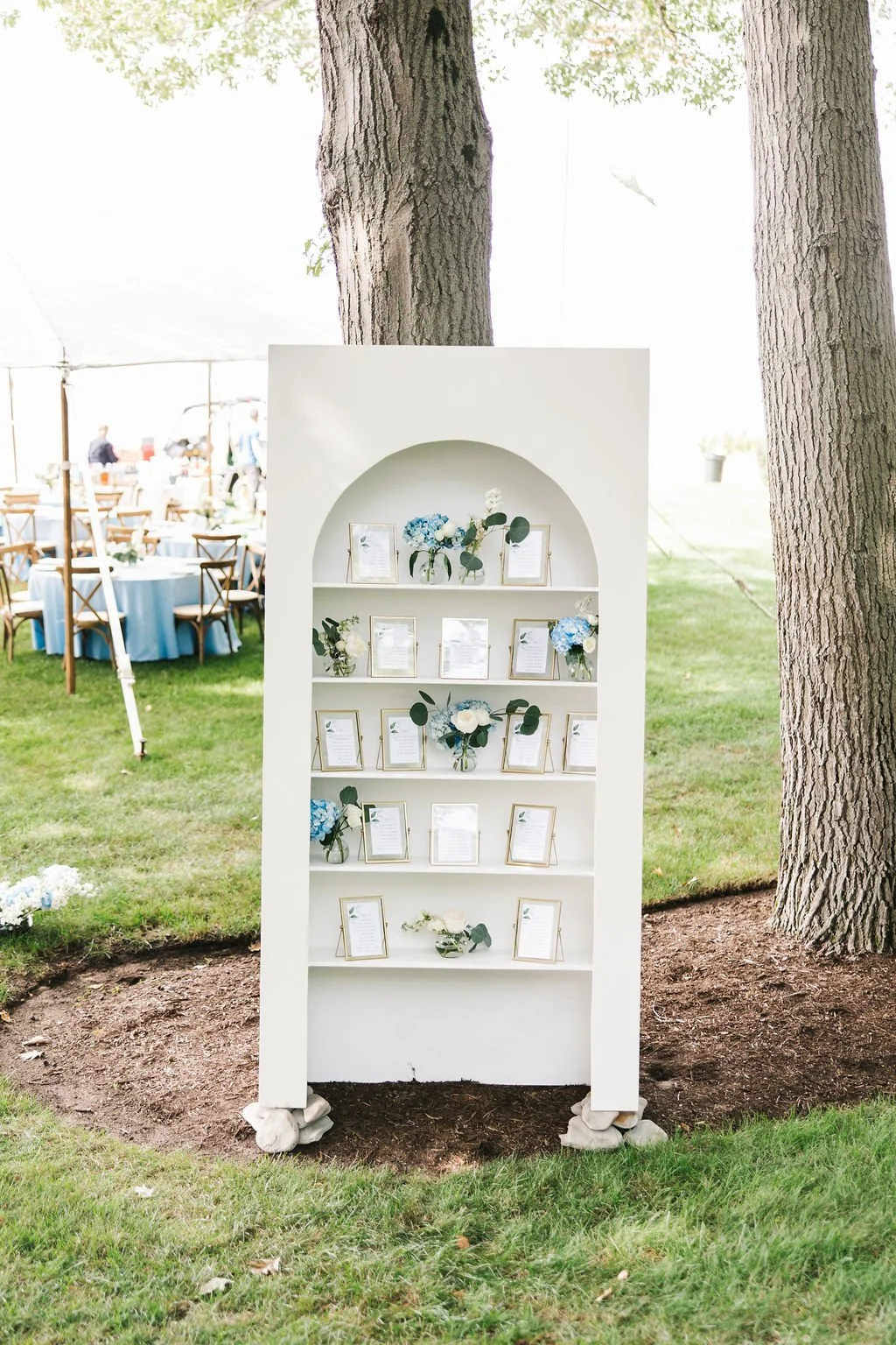 White display shelf with framed papers and blue and white flower arrangements, set outdoors under trees