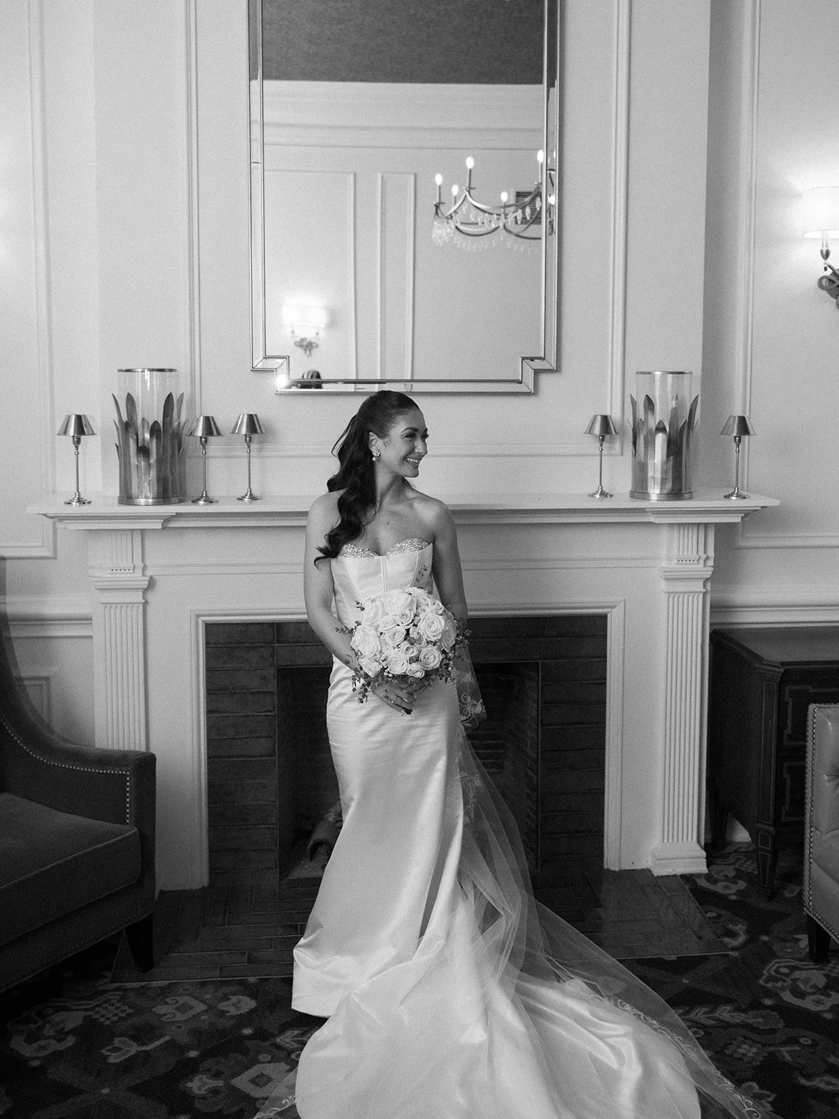 A smiling woman in a wedding dress holding a bouquet of roses, standing in a decorated room with a fireplace, mirror, and chandelier.