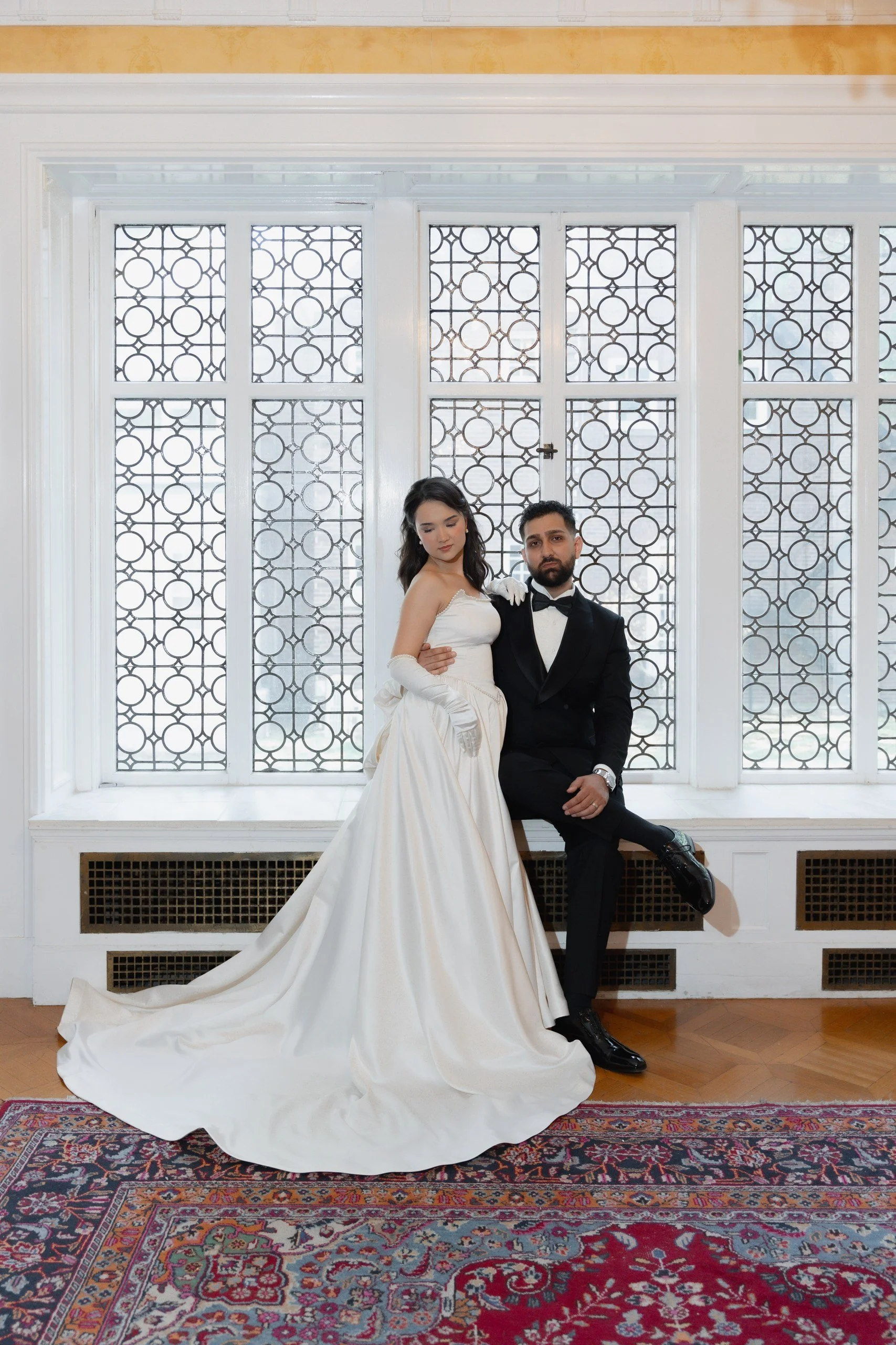 A bride and groom in formal attire standing in front of patterned glass windows. The bride is wearing a white wedding gown with long gloves, while the groom is dressed in a black tuxedo with a bow tie. They are posing indoors on a colorful patterned 