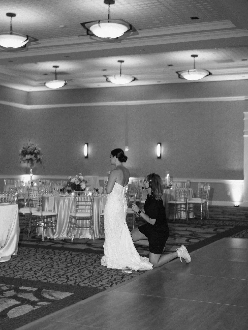 A bride in a wedding dress stands with her head bowed as a woman kneels in front of her, possibly proposing or preparing for a special moment, in a decorated banquet hall.