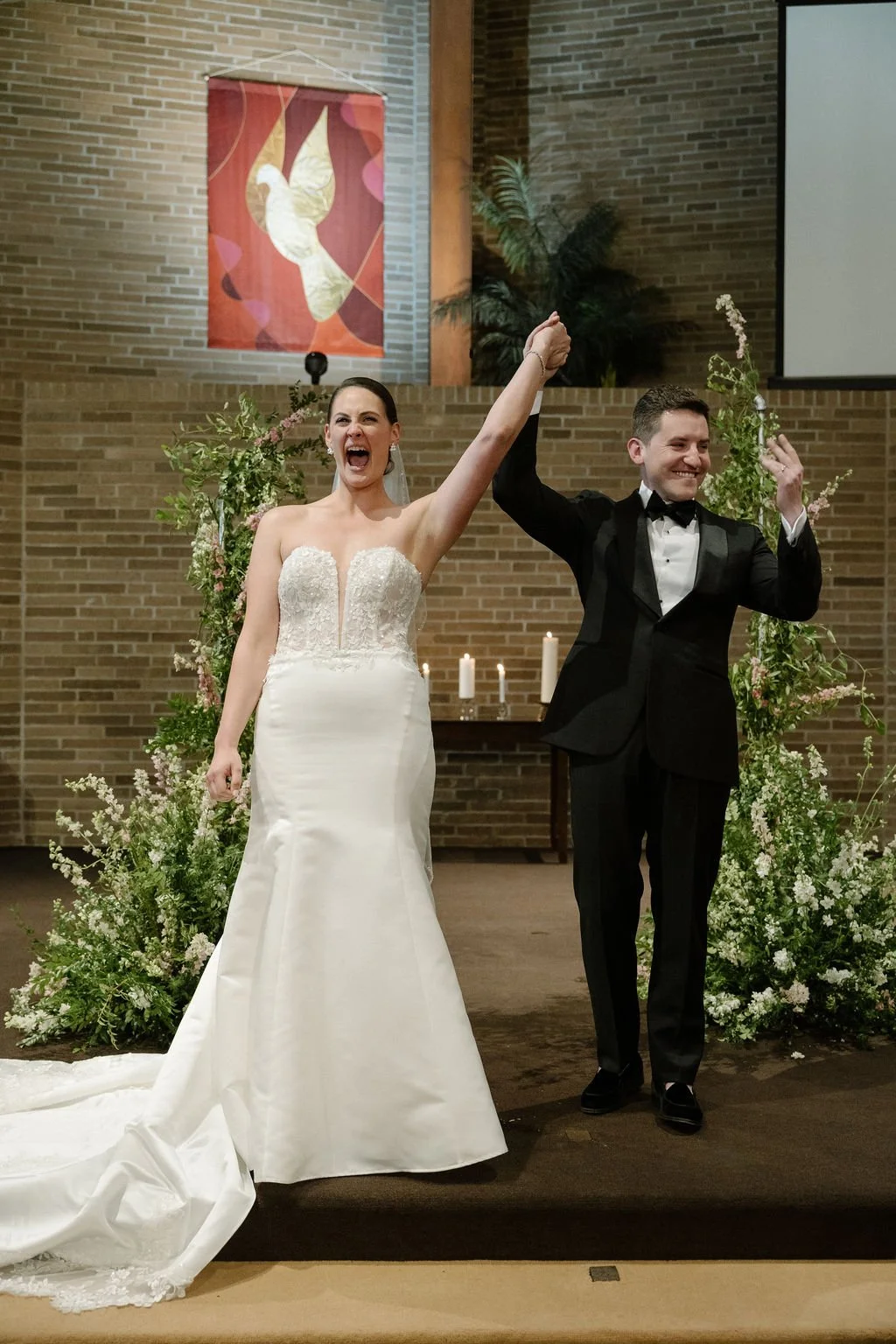 A bride and groom celebrating their wedding, holding hands raised in victory, standing in front of floral arrangements and candles in a church or chapel.