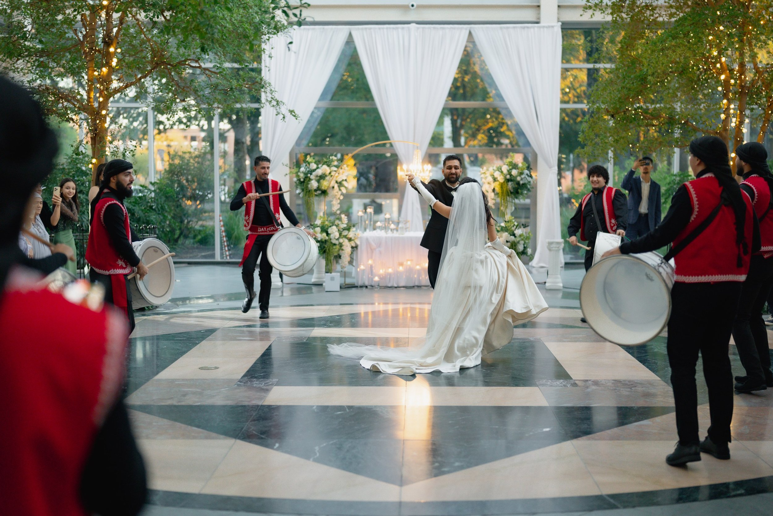A bride and groom are dancing at their wedding reception decorated with white flowers and drapes, with drummers playing in traditional attire around them.