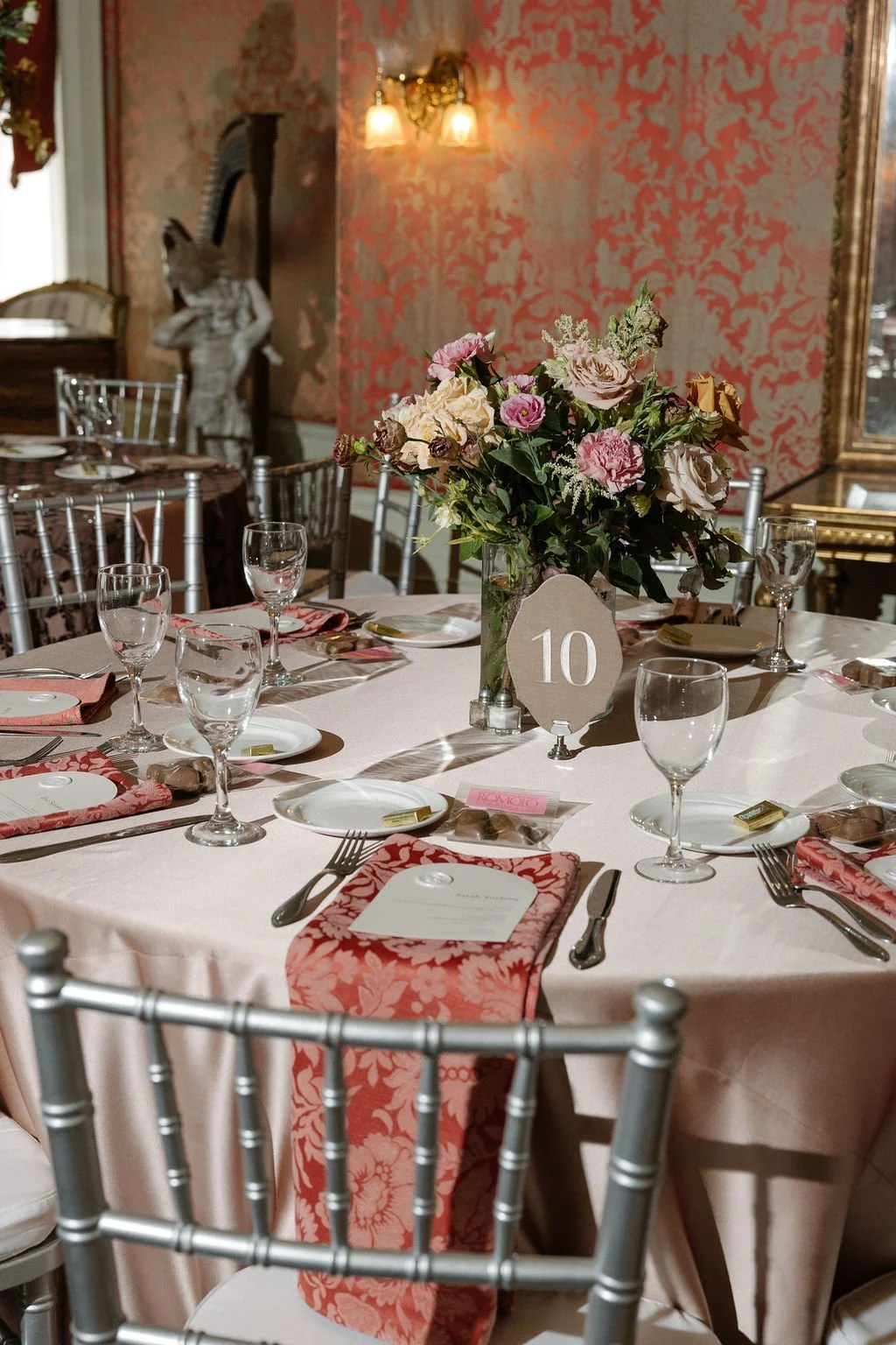 A decorated table set for a formal event with a floral centerpiece, glasses, plates, napkins, and utensils. The table has a pink tablecloth, pink floral napkins, and silver chairs. A table number '10' is displayed in the center.
