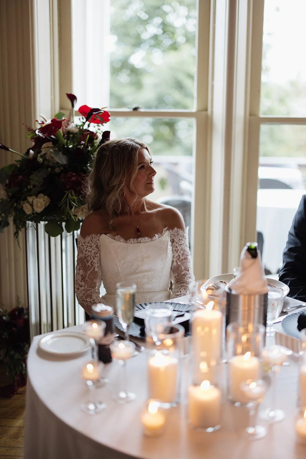 A woman in a white off-shoulder lace dress sitting at a table with candles during a wedding celebration.