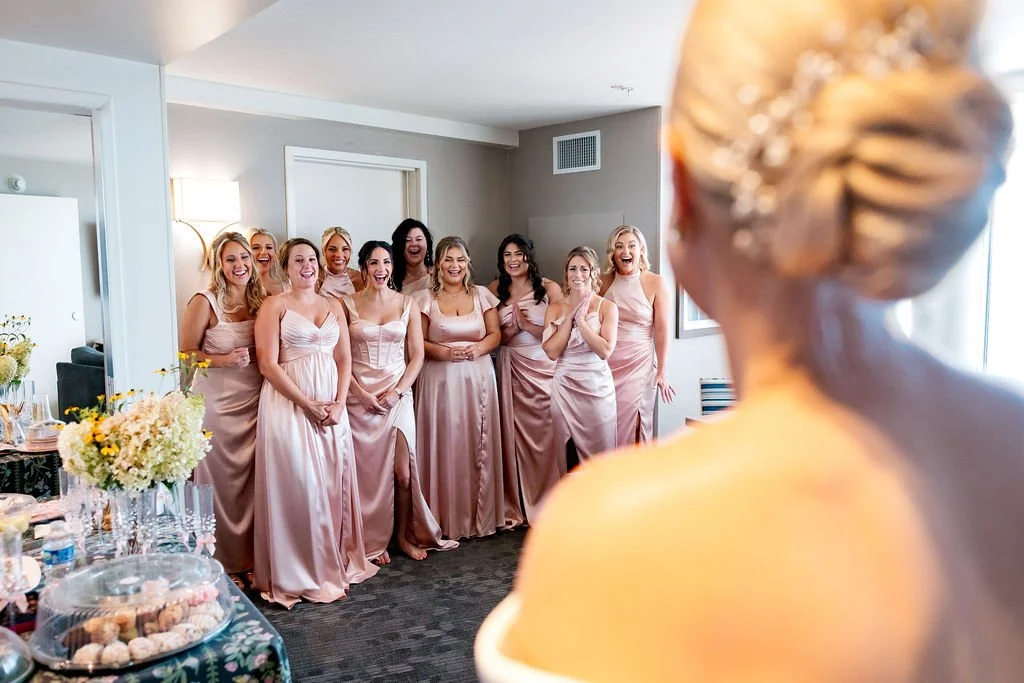A bride with blonde hair decorated with a hairpiece, seen from behind, facing a group of nine women in matching pink dresses. The women are smiling and appear happy and excited.