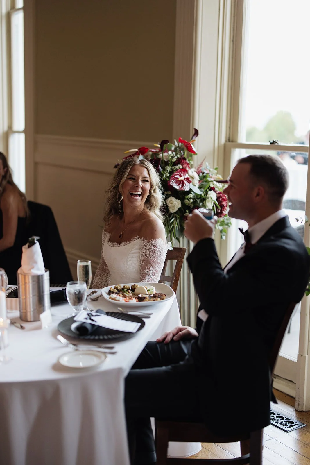 A joyful bride and groom at a wedding reception, sitting at a dinner table with a large flower arrangement and food on the table.