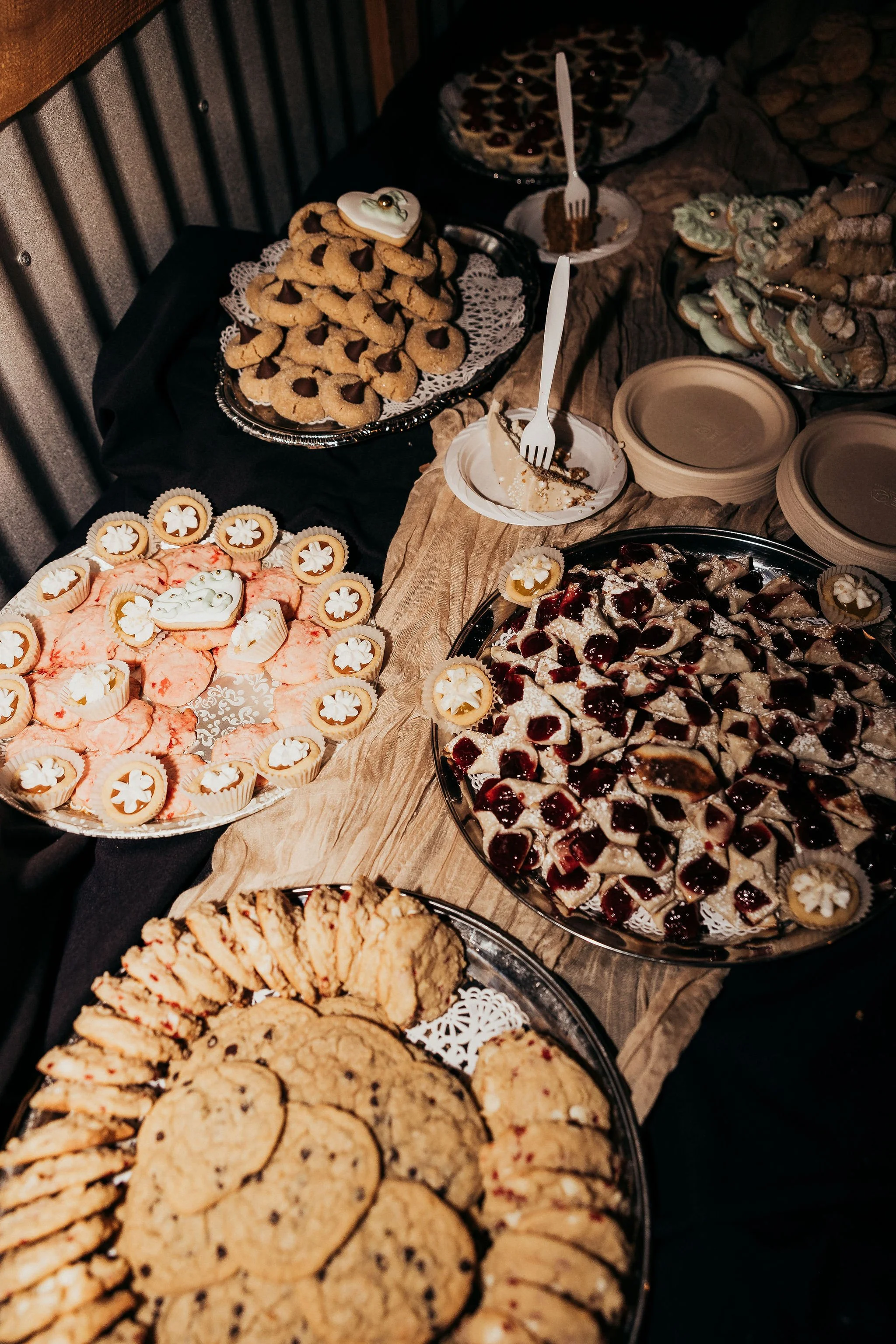 Assorted cookies, pastries, and desserts on a table with paper plates and forks, including chocolate chip cookies, thumbprint cookies with jam, mini tarts, and a pie with fruit topping.