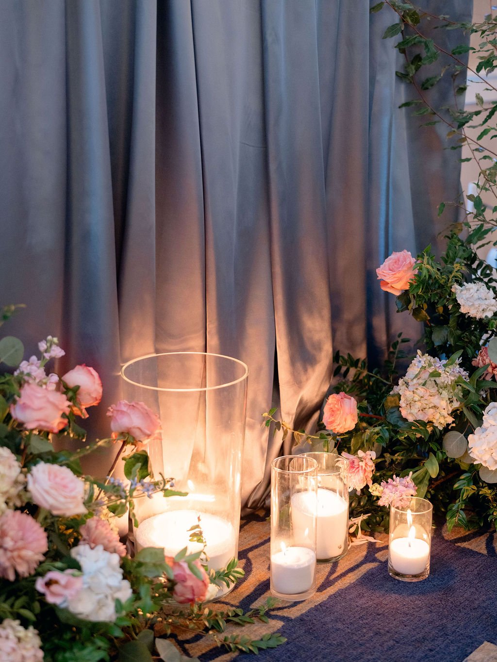 Several lit white candles in glass holders surrounded by pink and white flowers, set against a gray curtain backdrop.