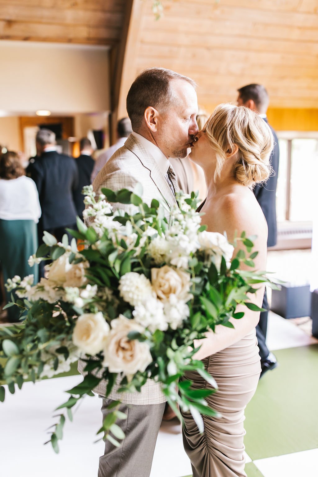 A couple kissing at a wedding reception with a bouquet of white flowers and greenery in the foreground.