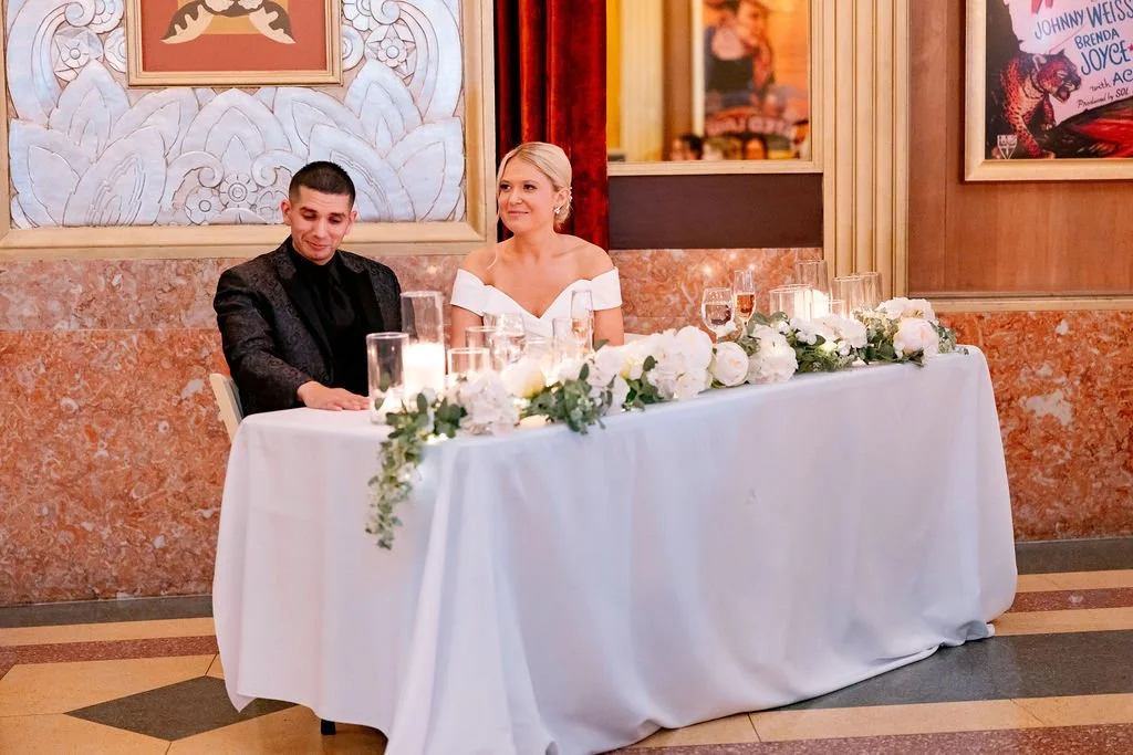 A bride and groom at a wedding reception sitting at a decorated table with white flowers and candles in a dimly lit room.