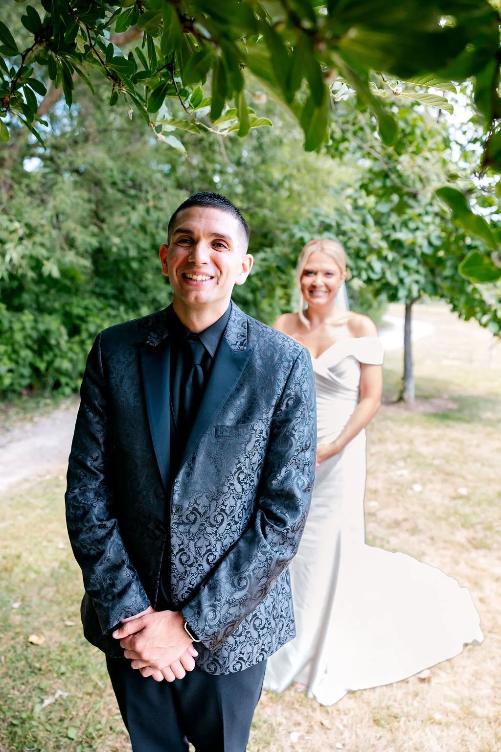 A groom in a dark patterned suit stands in the foreground, smiling, with a smiling bride in a white wedding dress in the background under green trees.