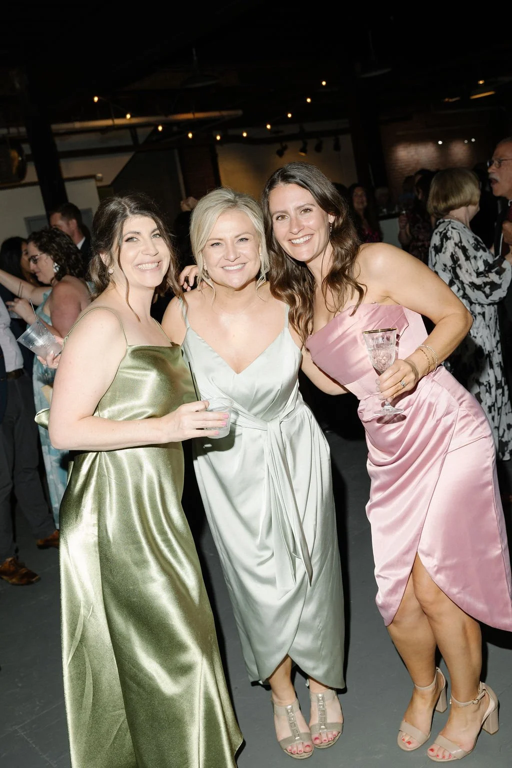 Three women in elegant dresses smiling and dancing at a party or wedding reception, holding drinks, in a dimly lit venue with other guests in the background.