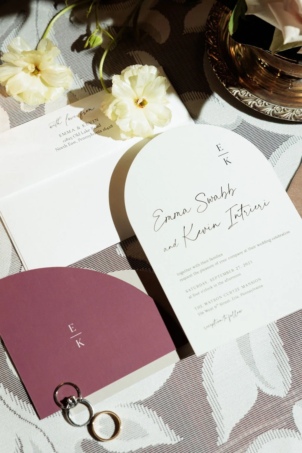 Wedding invitation suite with white and burgundy cards, cream flowers, and wedding rings on a patterned tablecloth.
