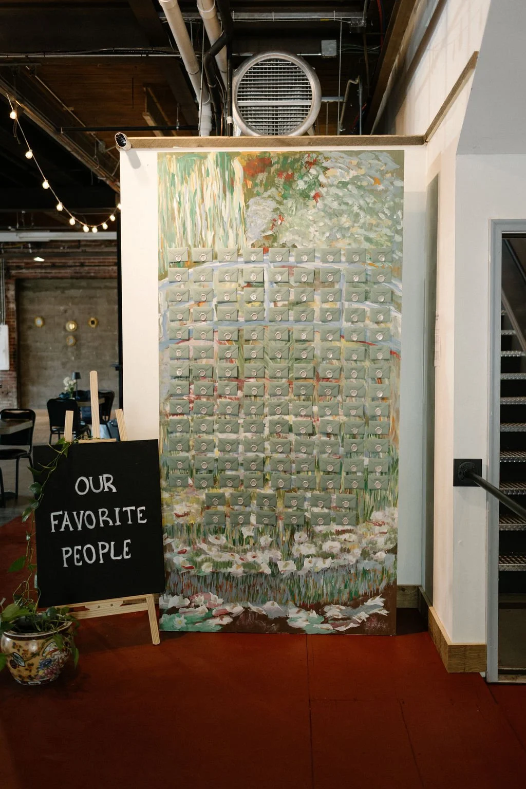 A painted backdrop resembling a grassy field with trees and bushes, covered with small, light green containers arranged in a grid, at an indoor venue. A blackboard sign reads 'Our Favorite People' with a small plant in front.