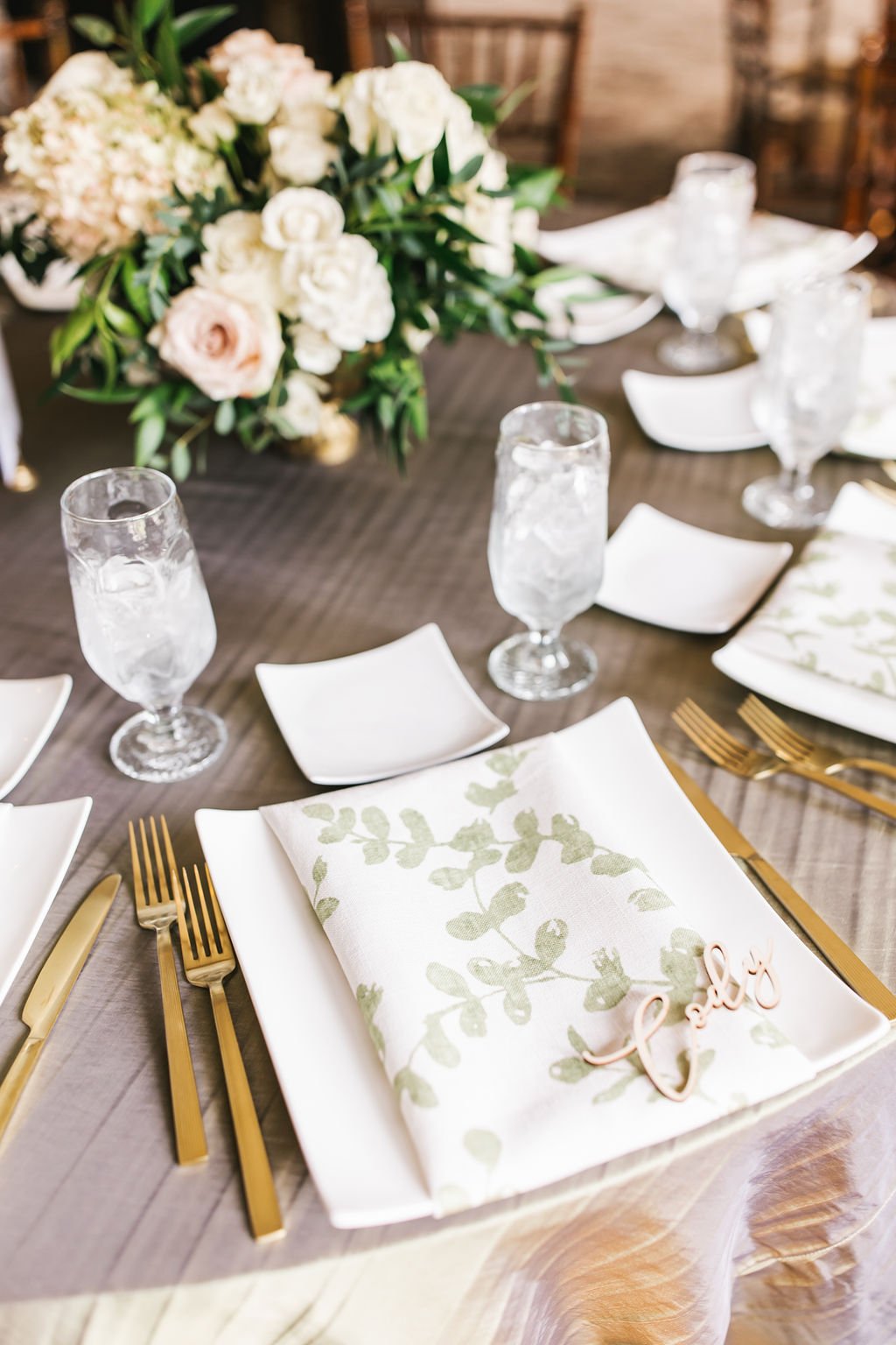 Elegant dining table set with gold utensils, white plates, napkins with green leaf patterns, glasses of water, and a floral centerpiece of white and light pink flowers with green leaves.